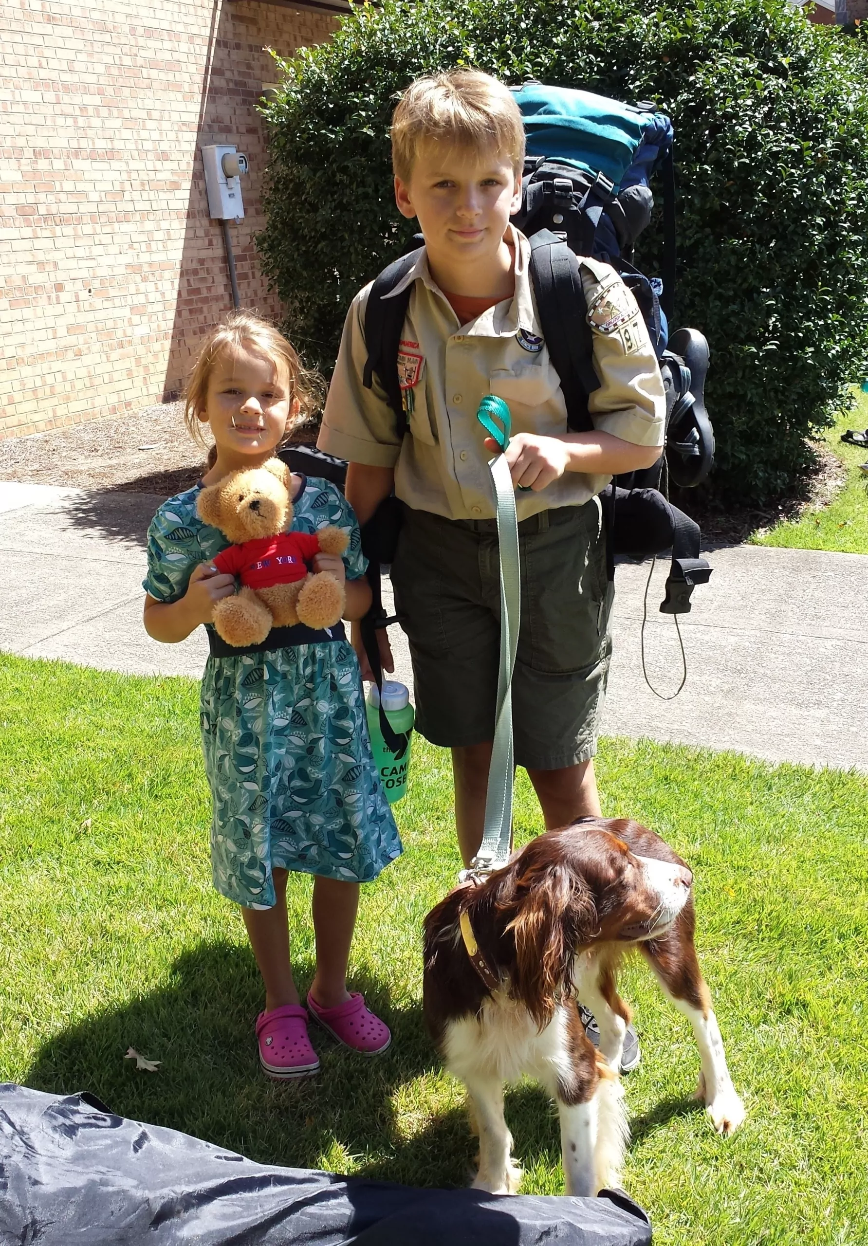 A young girl holding a stuffed bear smiles beside an older boy in a scouts uniform. The boy has a large bag of hiking gear on his back, holds a water bottle in one hand, and holds a leash in the other. A dog stands in front of them looking off to the side.