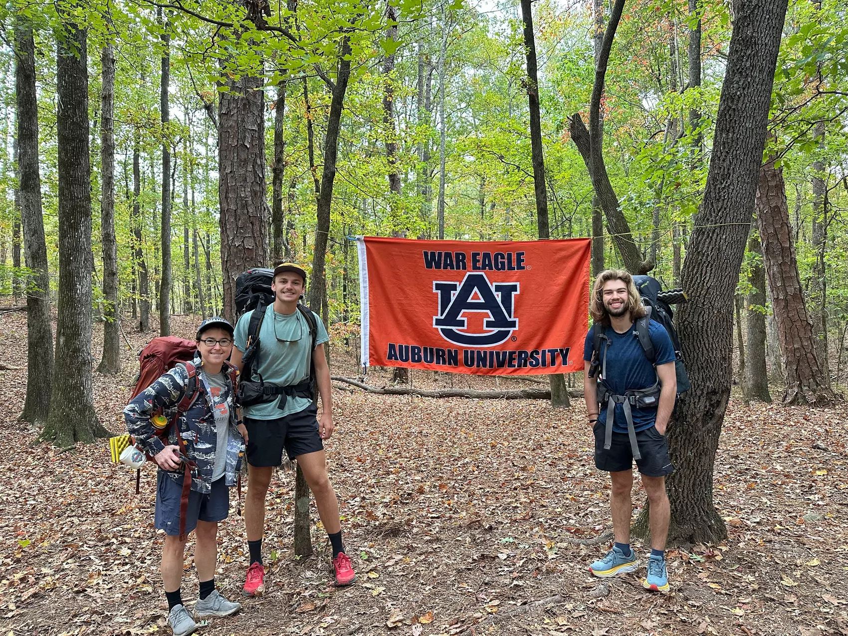 A young woman and two young men stand in the woods posing with a flag that says "War Eagle" and "Auburn University."